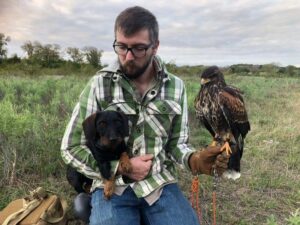 A man gently cradling a bird in one hand while his other hand rests on a loyal dog by his side.